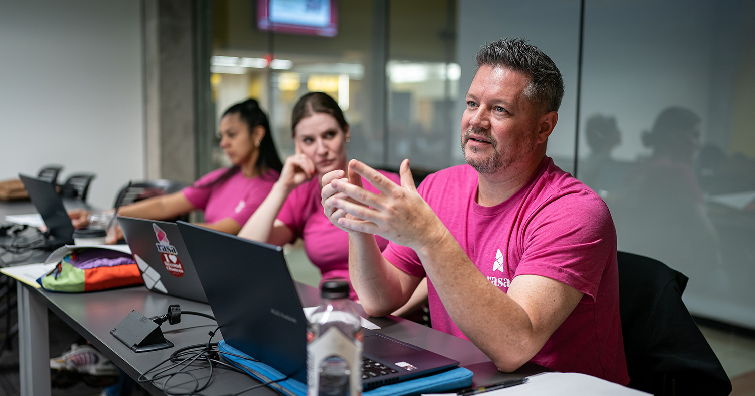 Photo of a man sitting in front of a laptop wearing a pink rasa shirt explaining