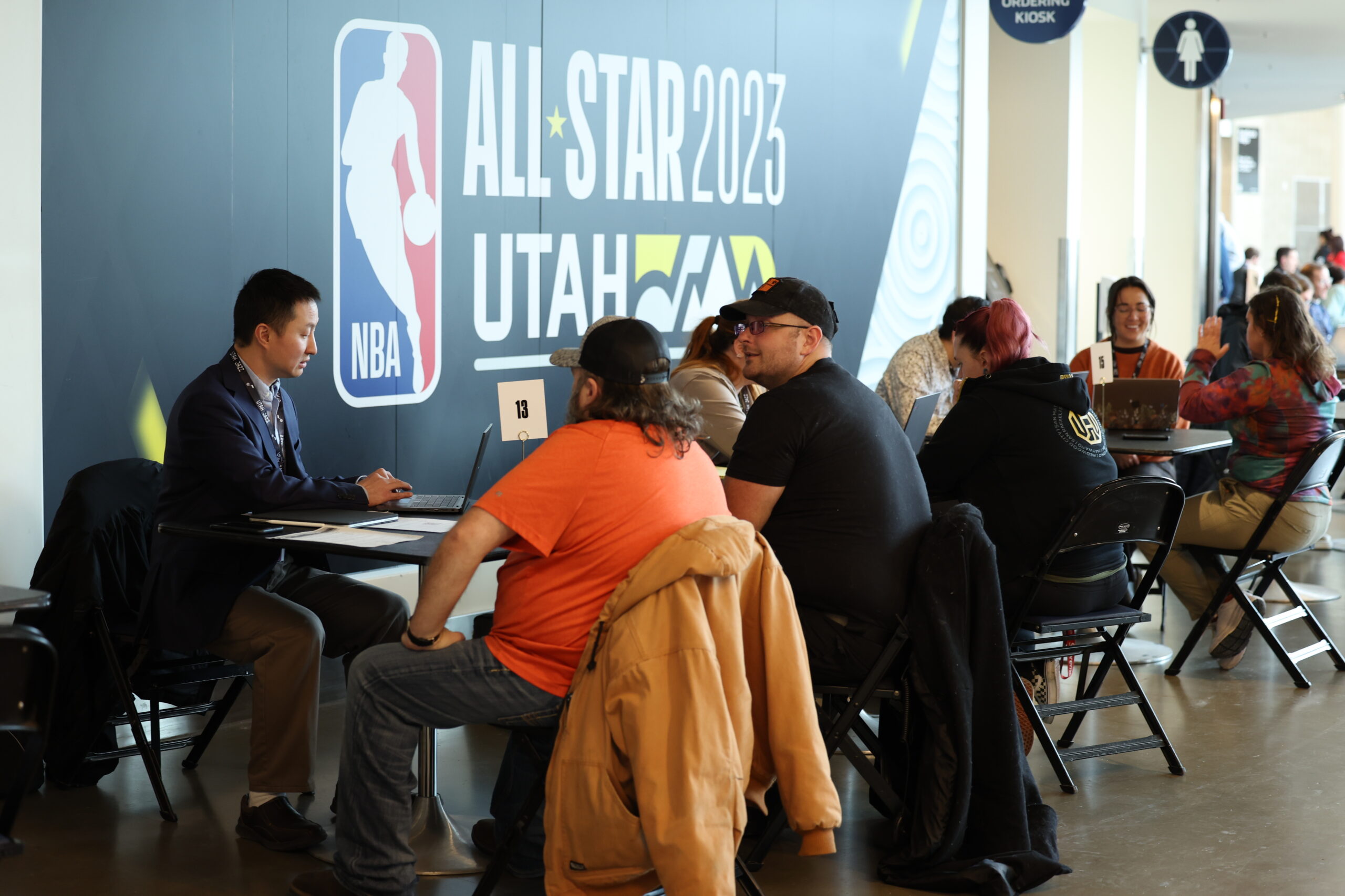 A group of people sitting at tables in a room with a large "NBA All-Star 2023 Utah" banner on the wall. A man in a suit is seated at a table with a laptop, engaging with attendees, while others are also interacting and using laptops.