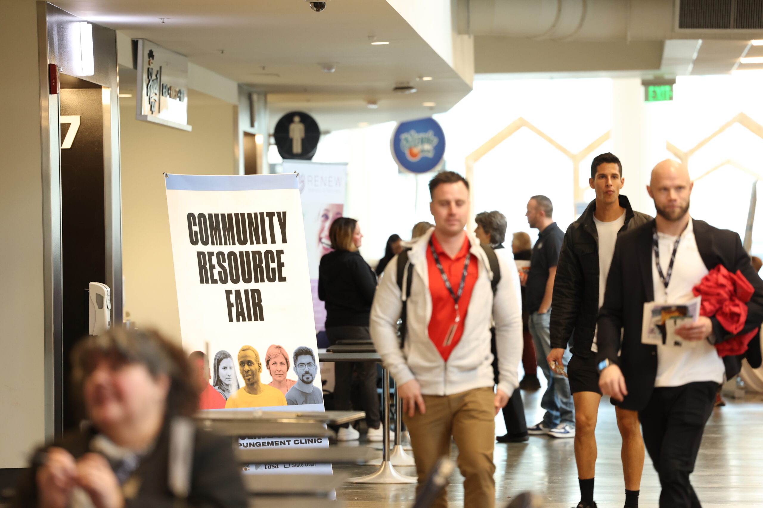 A hallway with people walking and mingling at a community resource fair. A large sign reading "Community Resource Fair" with photos of diverse individuals is prominently displayed in the foreground. People in the background are walking, talking, and holding materials.