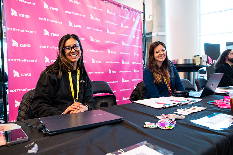 Two smiling women seated at a table with laptops and informational materials, set against a pink backdrop featuring the Rasa logo and #OffTheRecord.