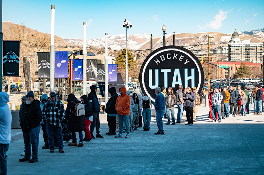 A long line of attendees waits outside the Utah Hockey sign at the Delta Center, with a scenic backdrop of mountains and the Utah State Capitol.