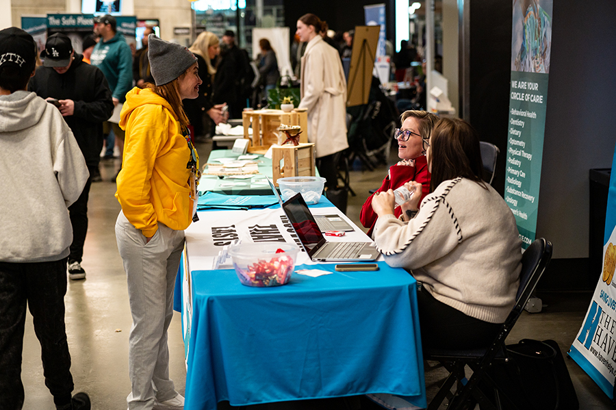 A woman in a yellow hoodie speaks with two representatives seated at a booth featuring informational materials, a laptop, and a welcoming display at a community event.