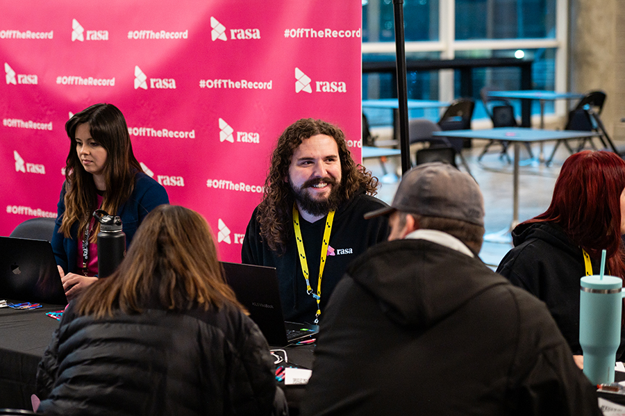 A man with curly hair and a cheerful smile engages with attendees at a table covered with laptops and materials, set against a pink Rasa-branded backdrop with #OffTheRecord.